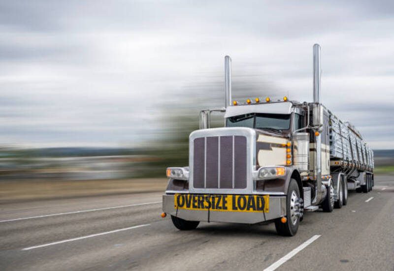 resized services 6 Classic dark long haul big rig bonnet semi truck tractor with oversize load sign on the front transporting fixed with slings lumber on the flat bed semi trailer running on the wide highway road